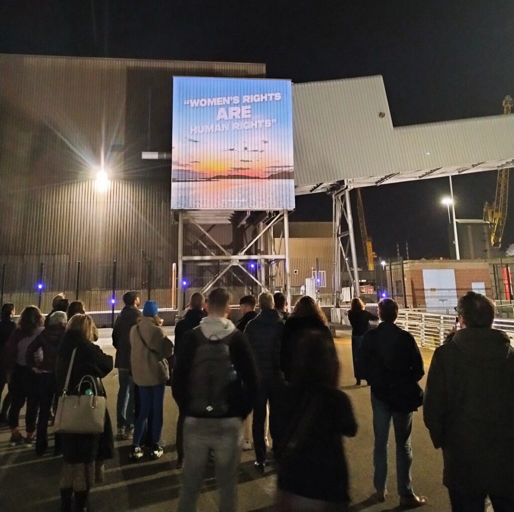 Group of people outdoors watching a presentation projected on a wall in an industrial setting. Text in the presentation says Women´s rights are human rights. 