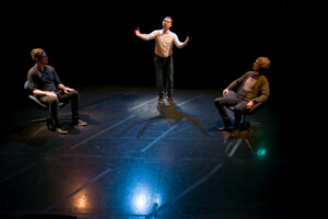 Three men on a stage and two of them are sitting, one is spreading hands wide.